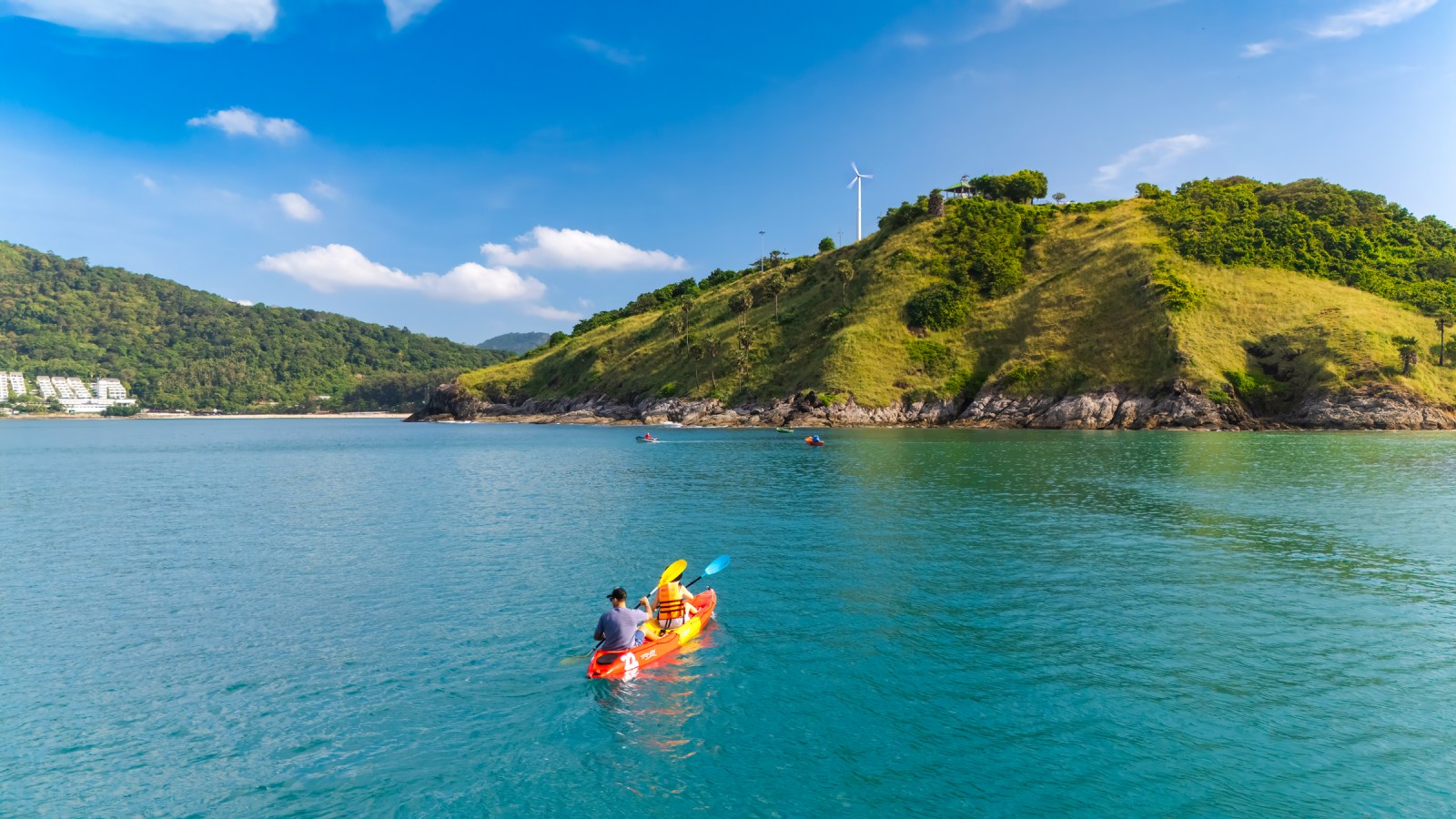 A person kayaking in a serene blue sea with a lush green hill in the background, featuring a wind turbine and scattered clouds in a clear sky.