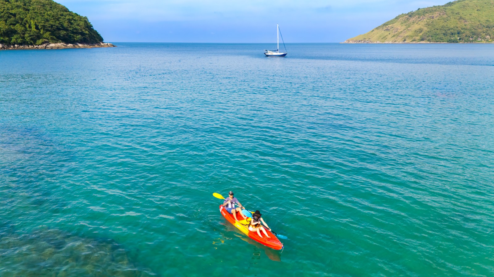 Two people kayaking on clear turquoise waters with a sailboat in the background.
