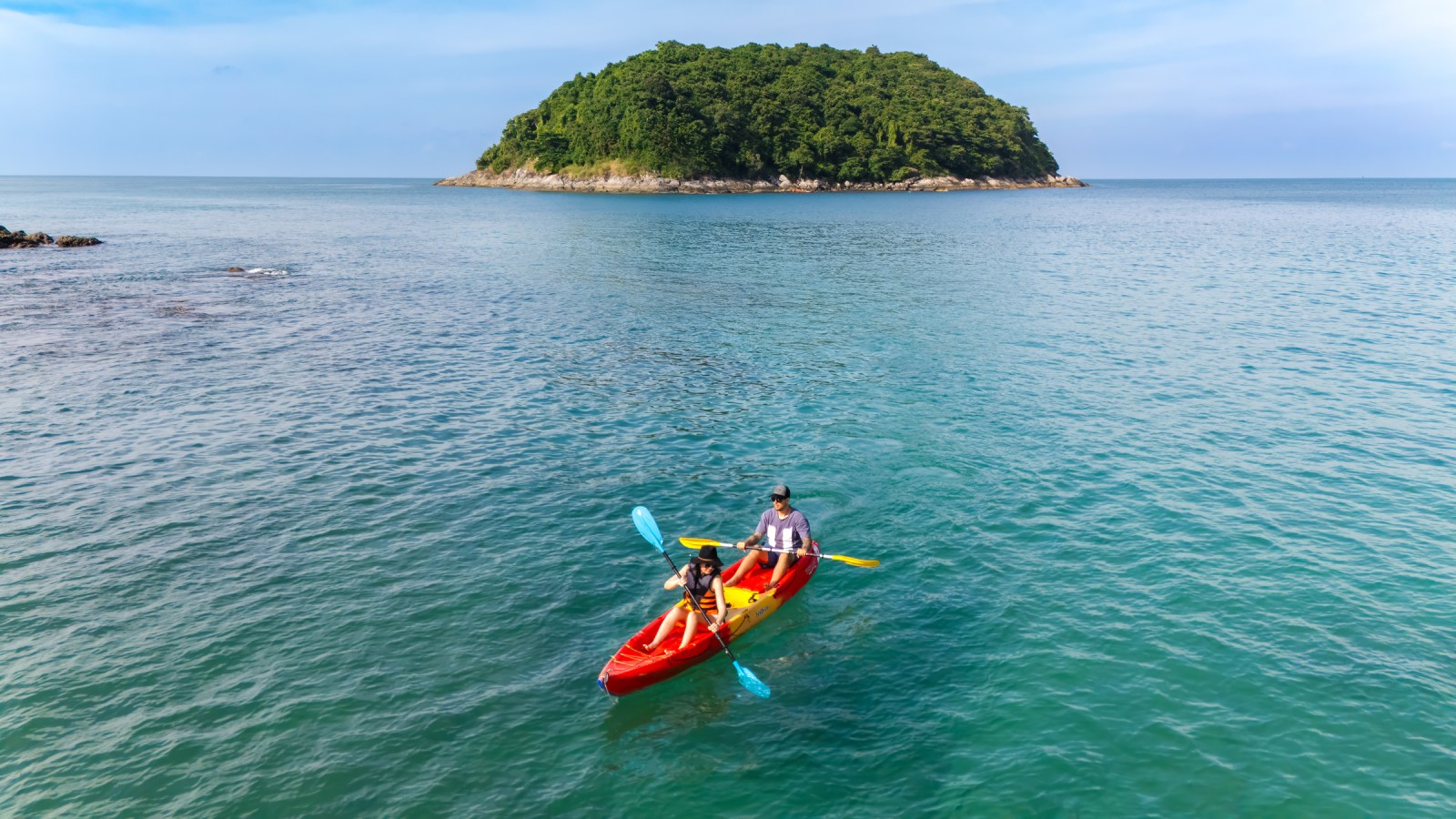 A canoe with two people paddling in clear blue water, with a small, lush green island in the background.