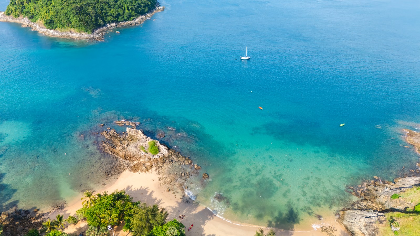 Aerial view of a tranquil beach with clear blue water, rocky shoreline, and greenery surrounding the area.