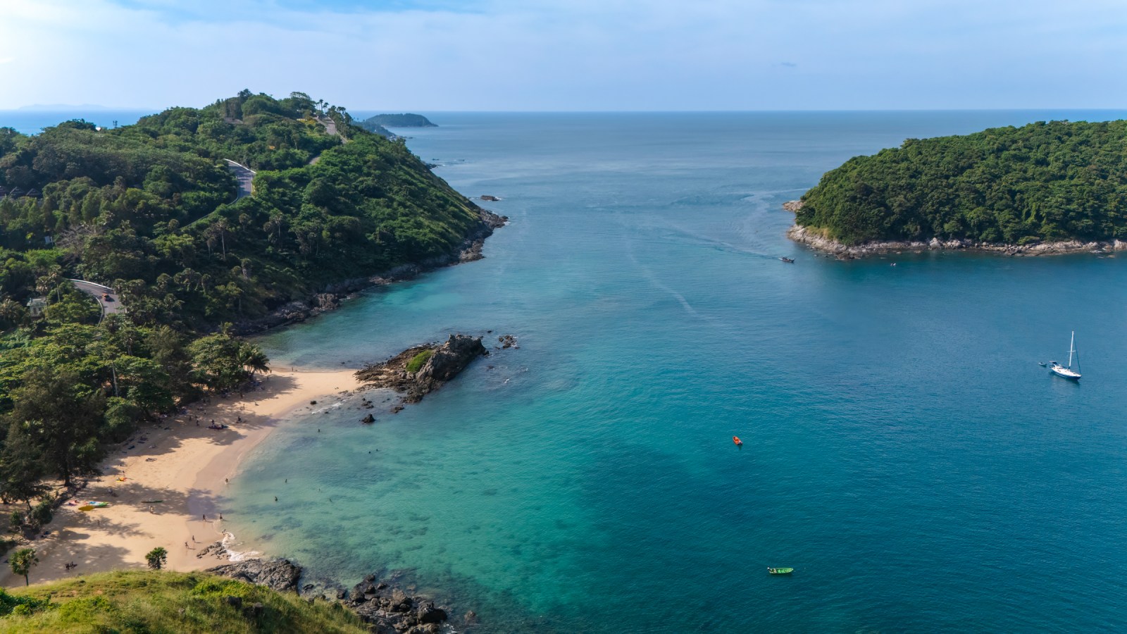 Aerial view of a beautiful beach surrounded by lush green hills and clear blue water, with a few boats visible on the water.
