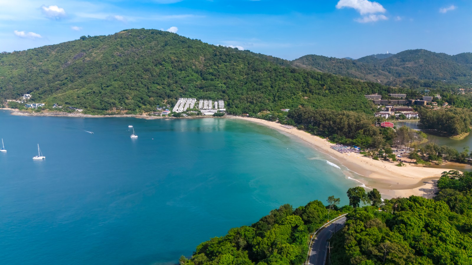 Aerial view of a serene beach surrounded by lush green hills and clear blue water, featuring a few boats near the shore.