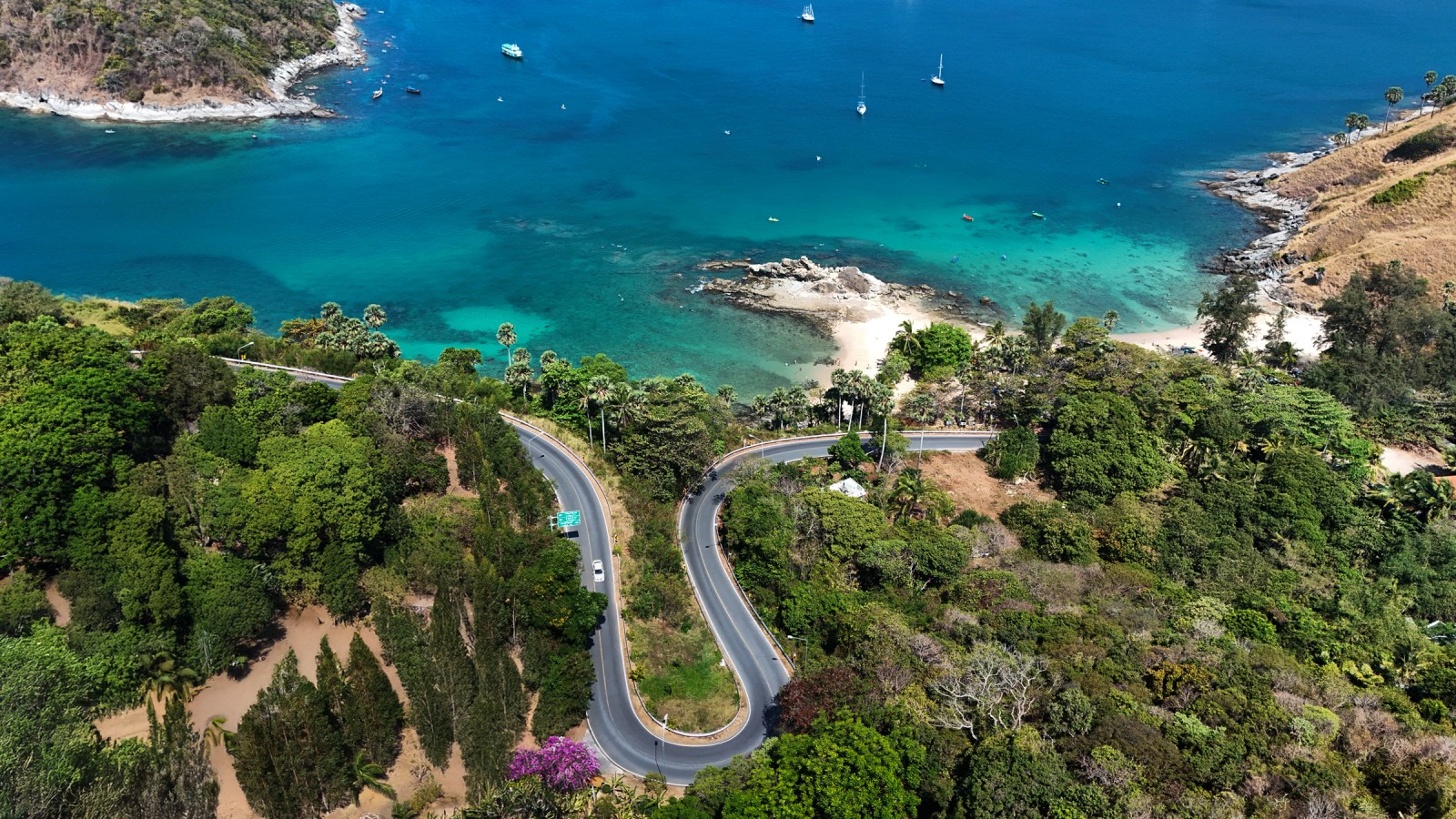 Aerial view of a winding road surrounded by lush greenery leading to a turquoise sea with boats anchored near a beach.
