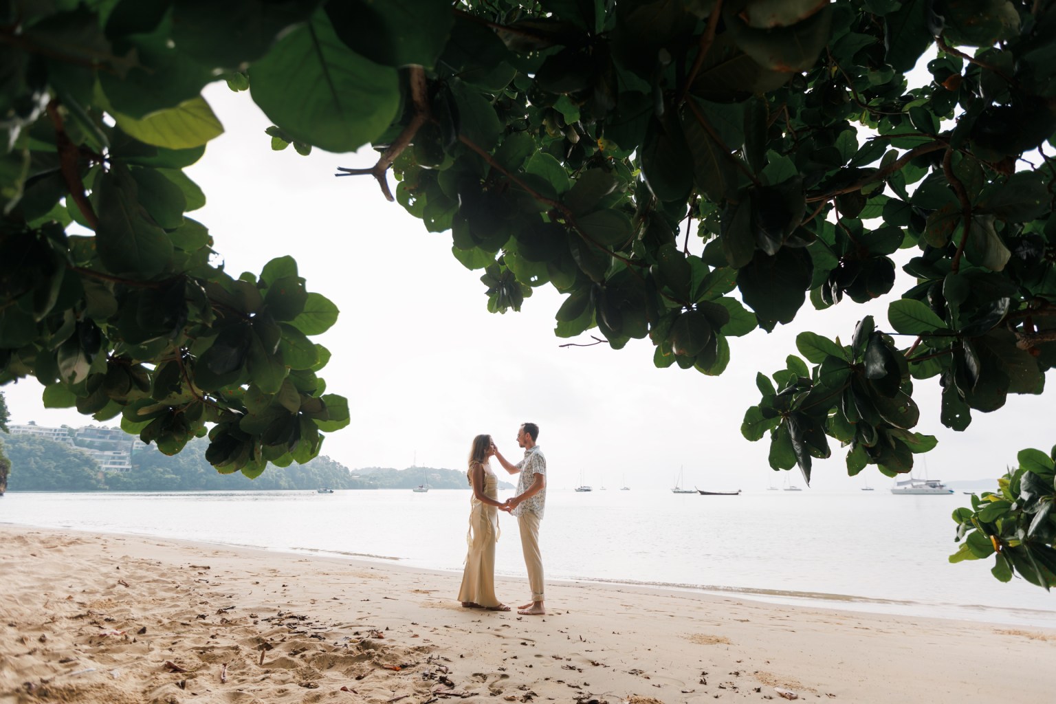 A couple standing hand in hand on a beach, framed by lush green leaves in the foreground, with a serene ocean view in the background.