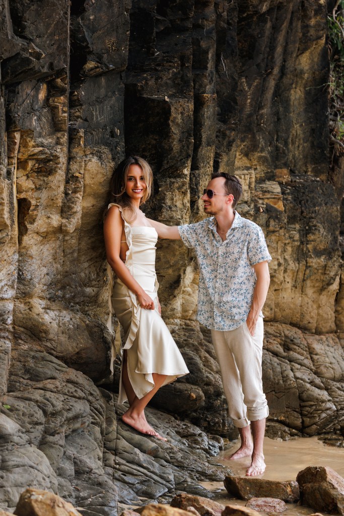 A couple standing on a rocky beach, the woman in a white dress and the man in a floral shirt, smiling and posing for the photo.