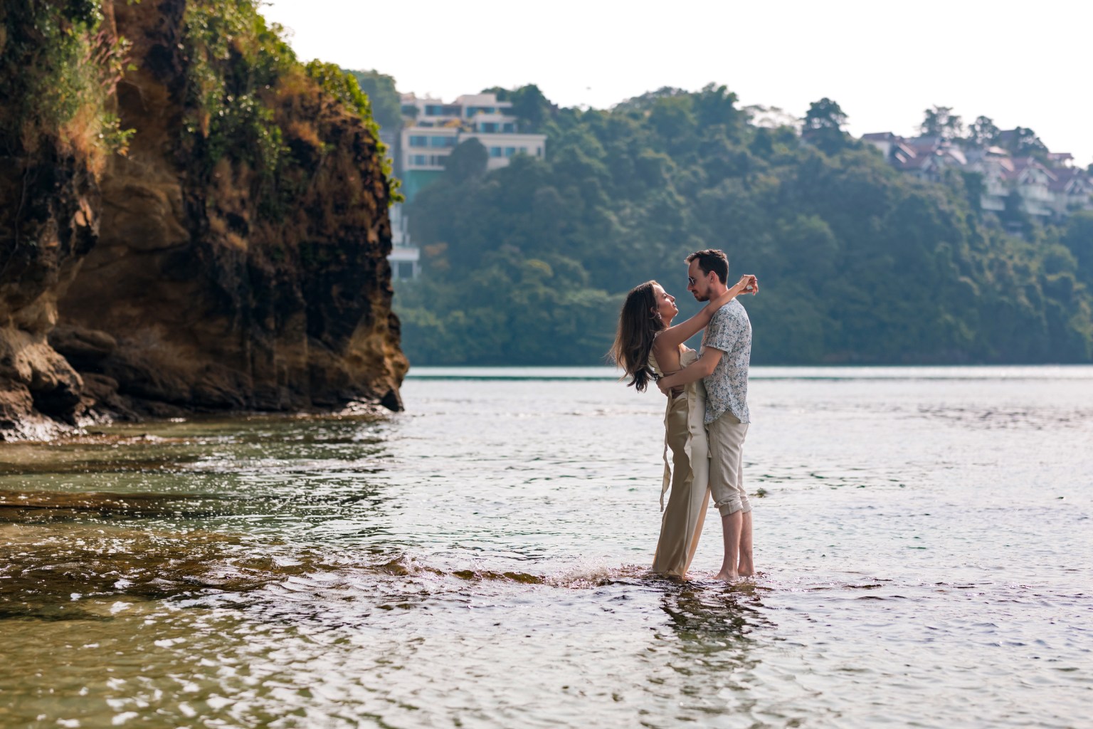 A couple standing in shallow water, embracing each other amidst a scenic backdrop of cliffs and greenery.