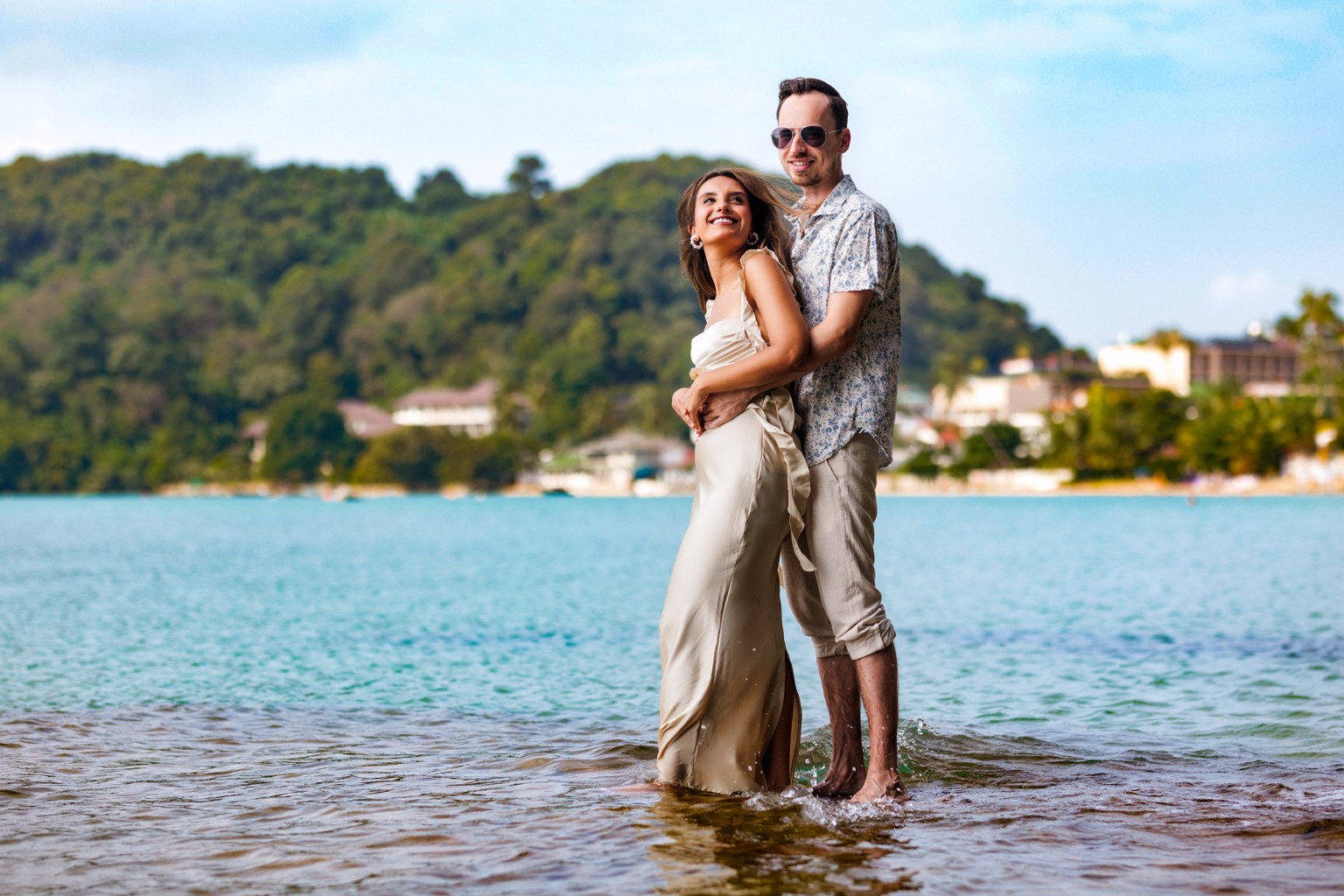 A couple standing in shallow water, smiling and embracing, with a scenic beach and hillside in the background.