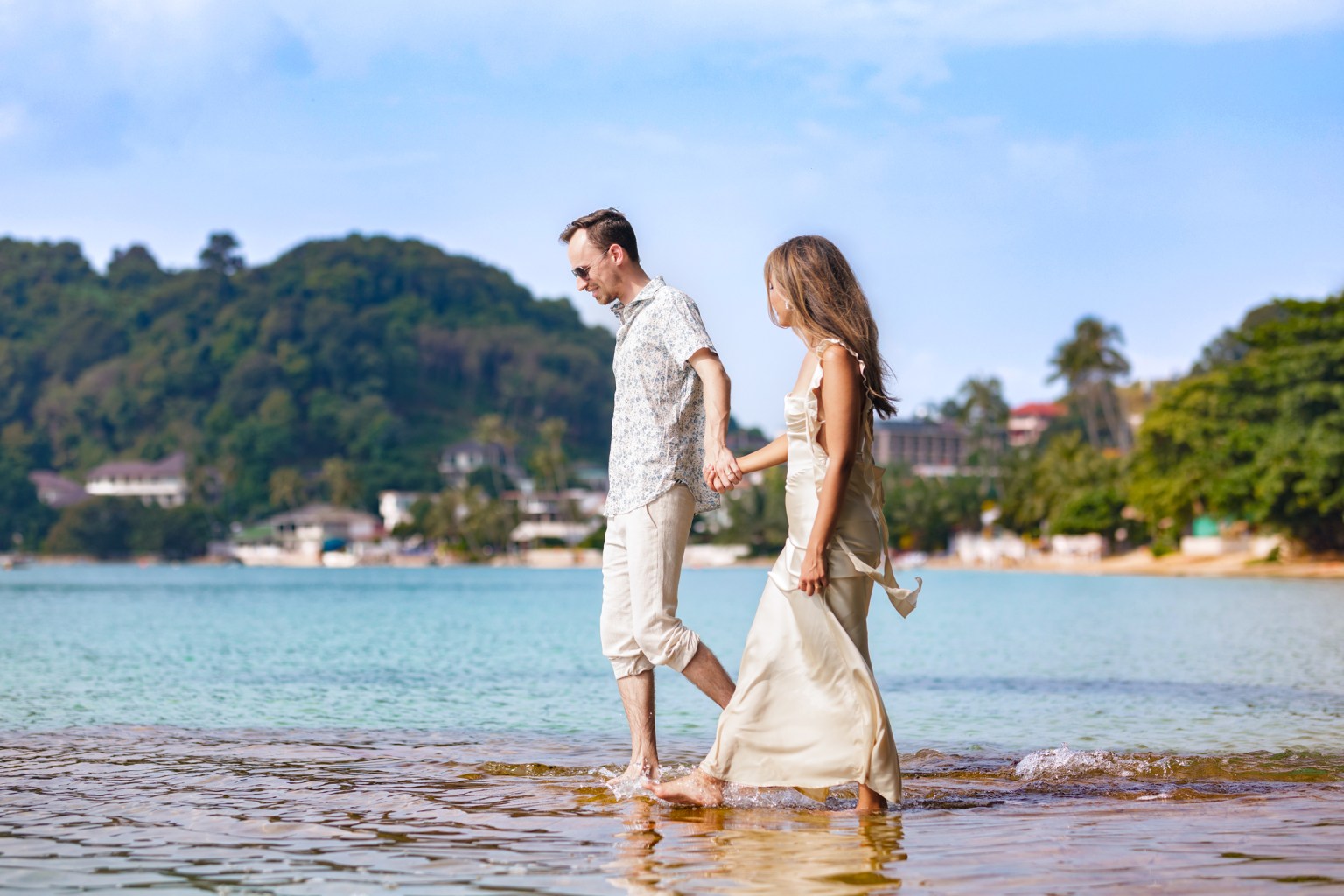 A man and a woman walking hand in hand along a beach, with gentle waves lapping at their feet and a scenic landscape in the background.