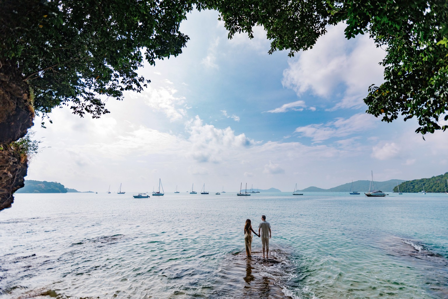 A couple holding hands standing in shallow water at Ao Yon Beach, surrounded by lush greenery and sailboats in the background.