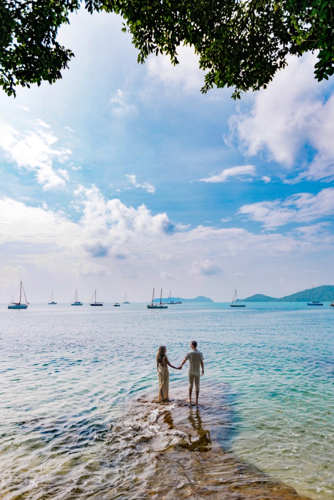 A couple holding hands while standing in shallow water, with sailboats in the background and a bright blue sky overhead.