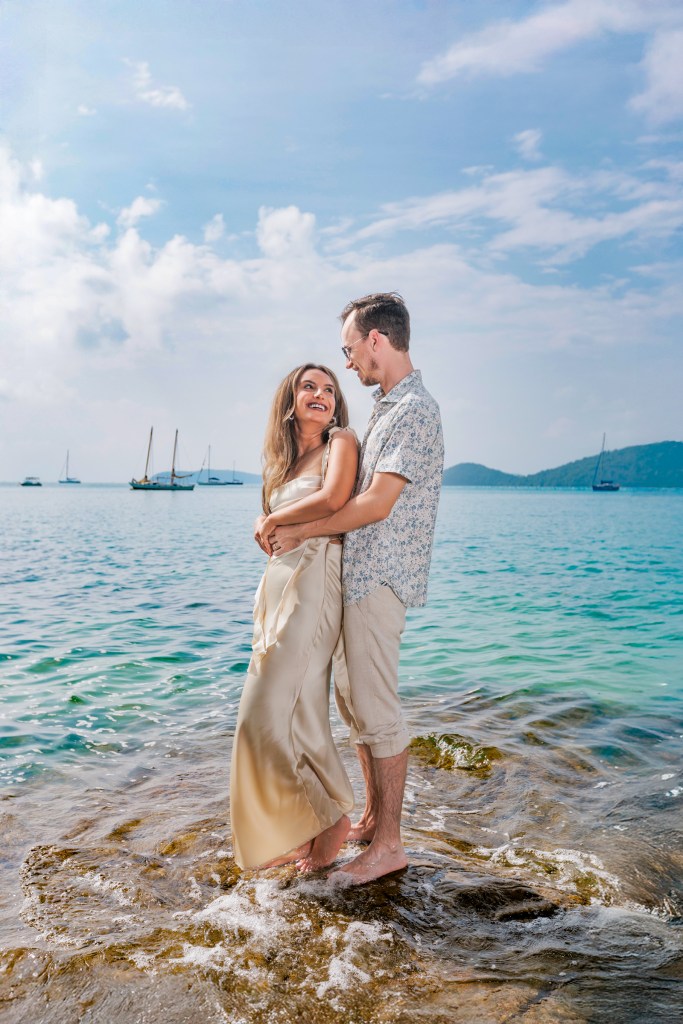 A couple standing on a rocky shore in the ocean, embracing each other, with a backdrop of sailboats and a clear blue sky.