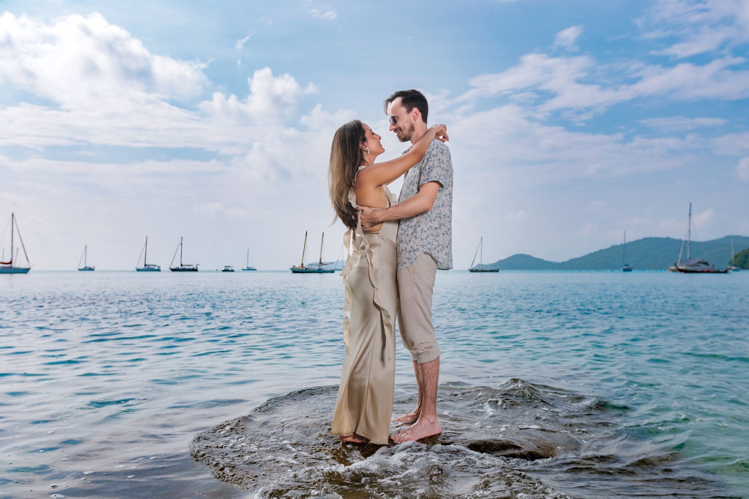 A couple embracing on a rock in the water, with sailboats in the background and a cloudy sky.