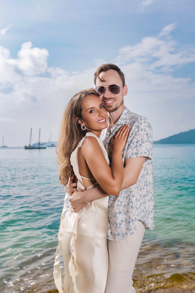 A couple embracing near the water, with a scenic view of the ocean and distant boats in the background. The woman is wearing a light-colored dress and the man is dressed in a patterned shirt and sunglasses.