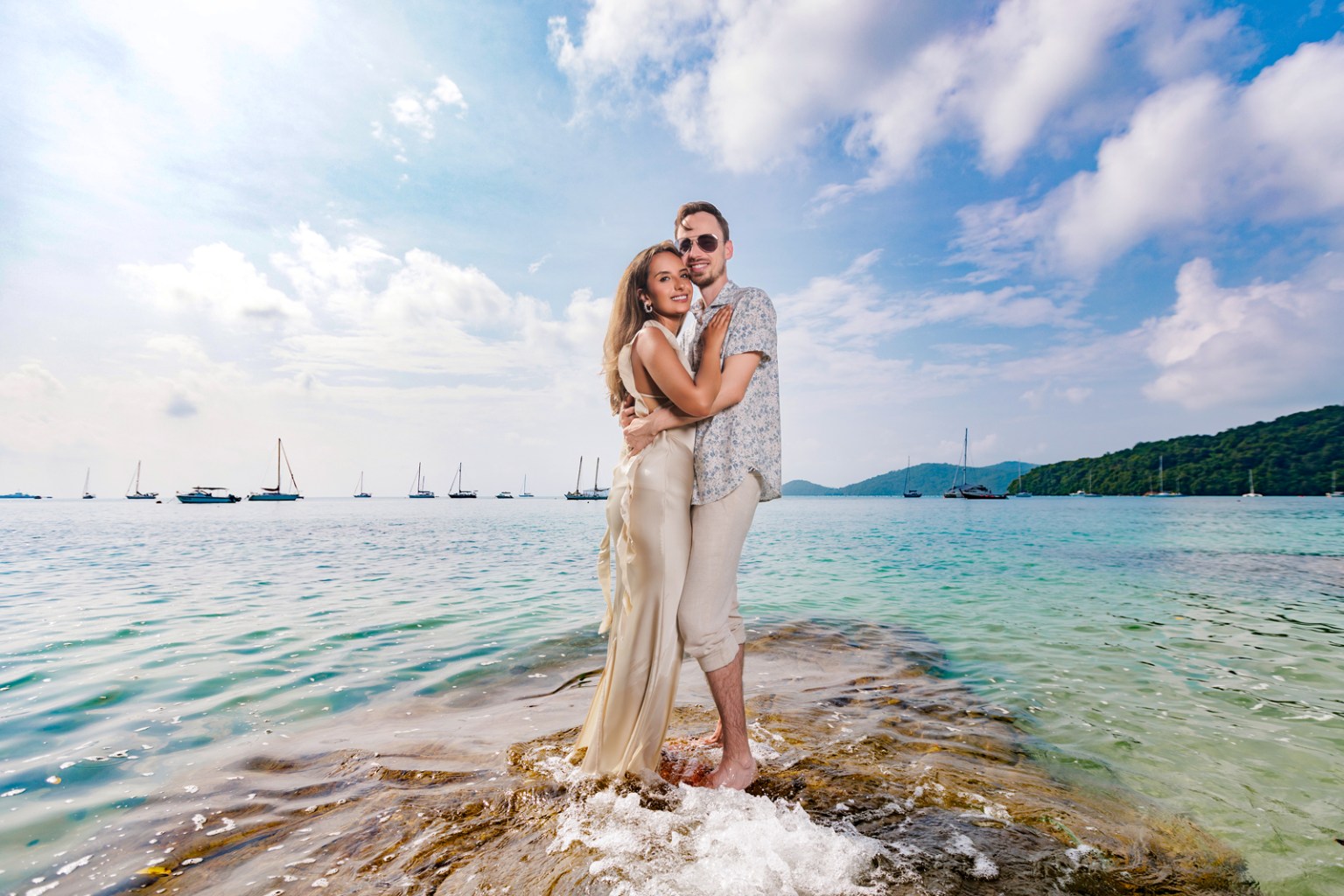 A couple standing on a rock by the shore, embracing each other with a scenic view of the beach and boats in the background.
