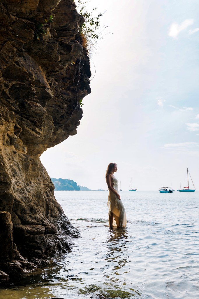 A woman standing in shallow water near a rocky shoreline, with boats in the distance under a cloudy sky.