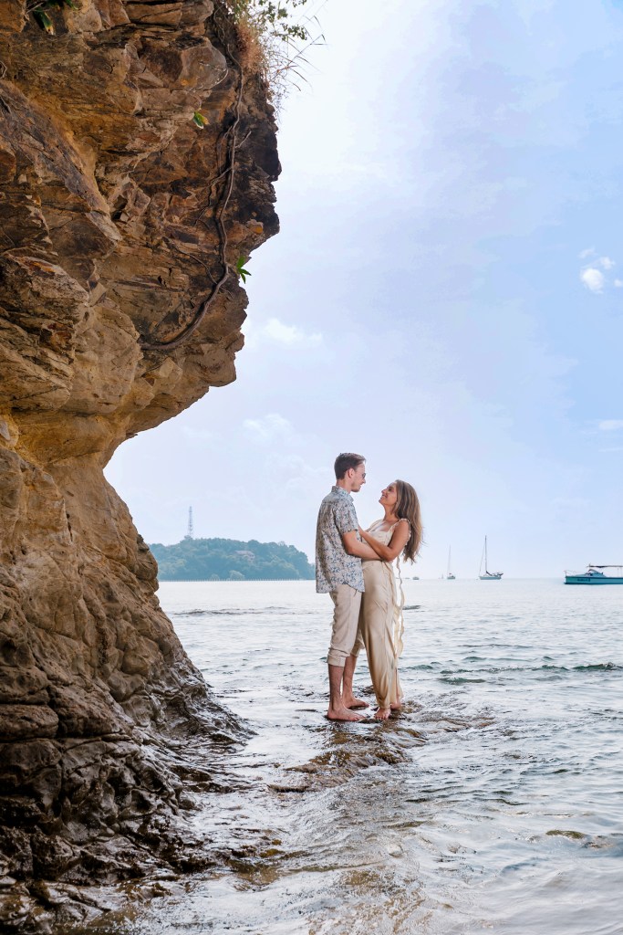 A couple standing by the shore, embracing each other with a rocky cliff in the background and a scenic ocean view.