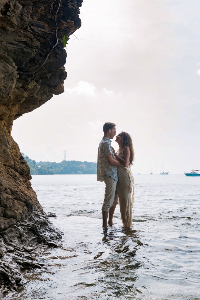A couple standing in shallow water near a rocky shore, embracing each other, with boats visible in the background and a cloudy sky above.