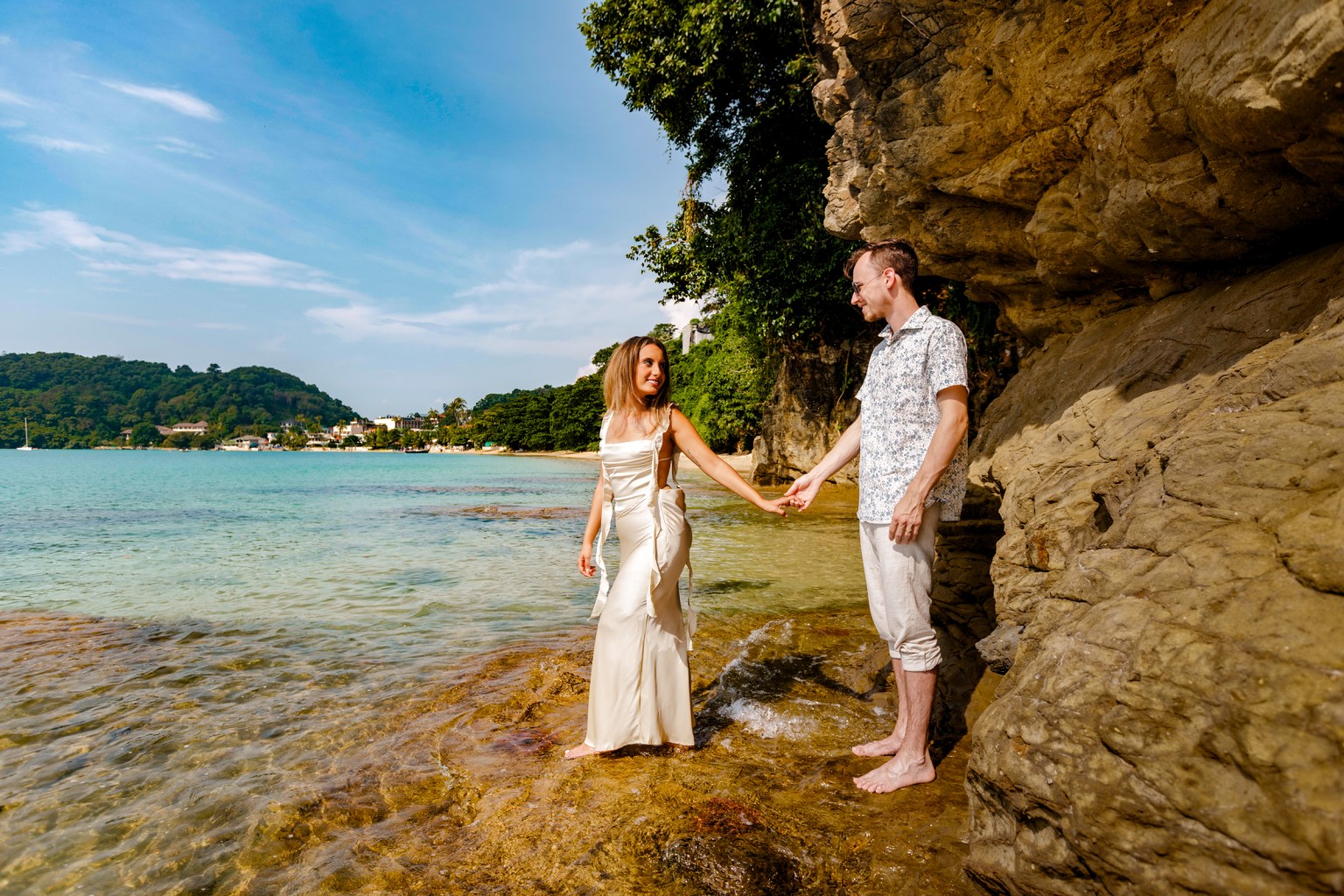 A couple holding hands on a rocky beach by the clear water, with trees and buildings in the background.