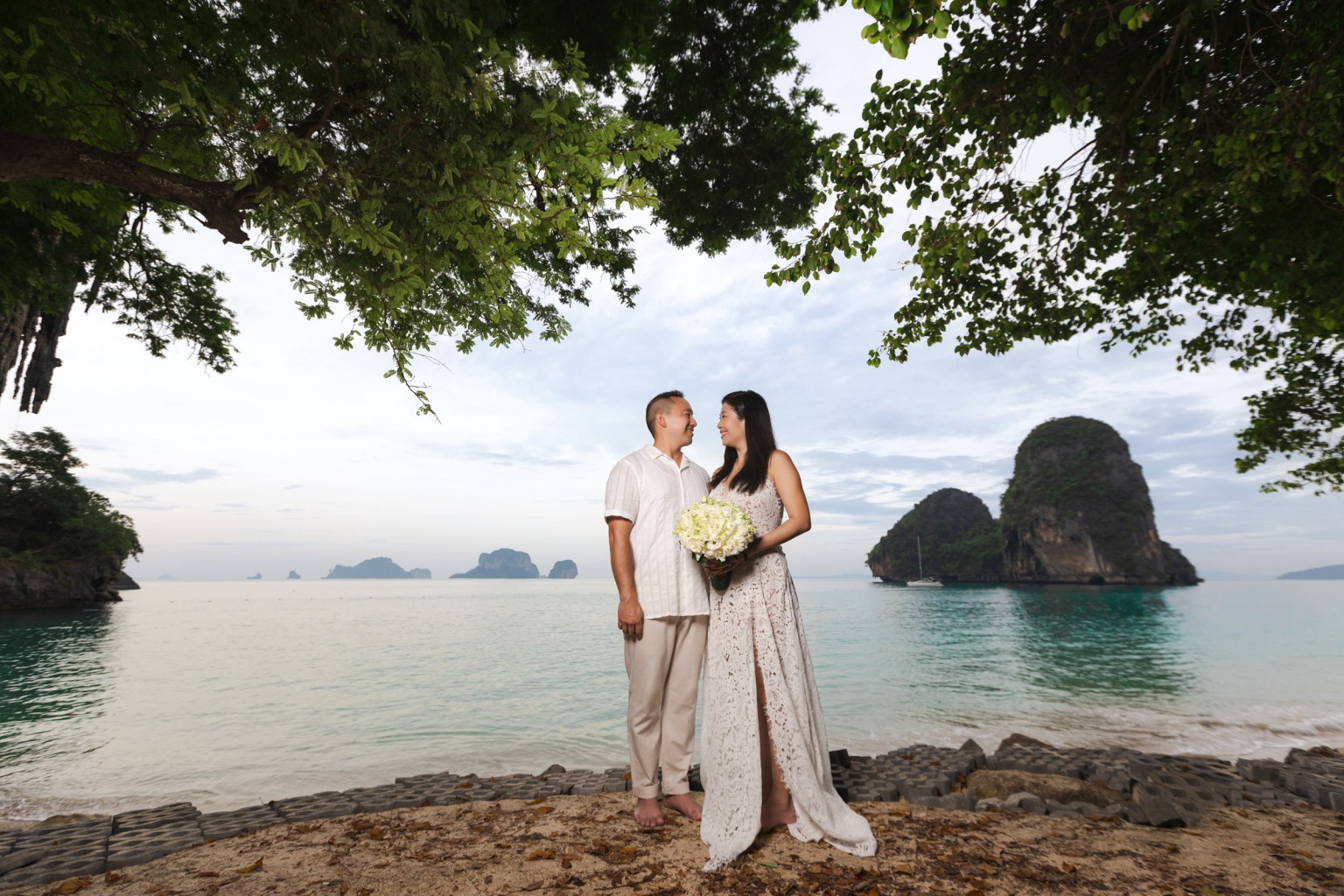 Couple standing on a beach with a bouquet, looking at each other, surrounded by trees and rocky formations in the background.