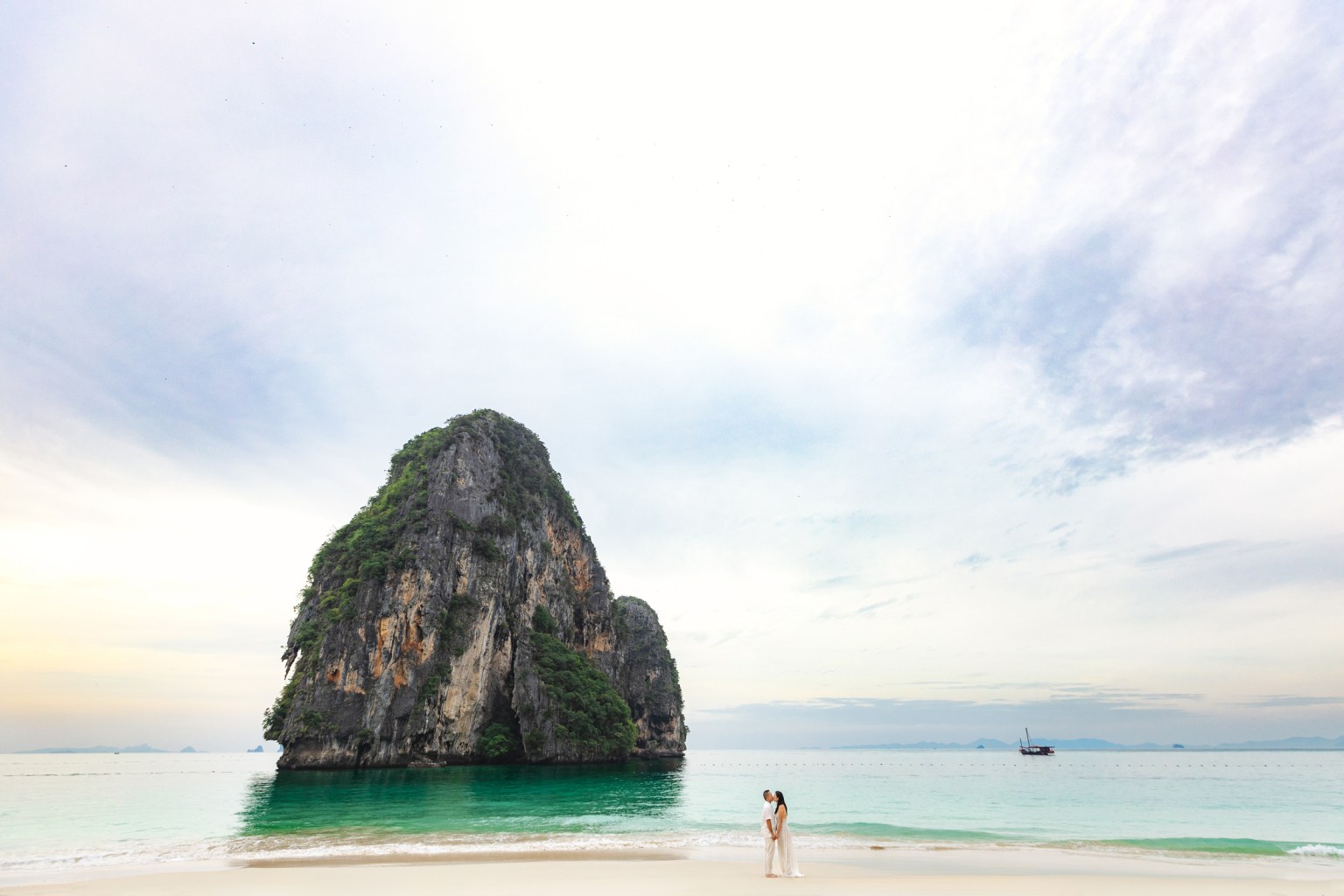 Couple embracing on a beach with a large rocky formation in the background and calm turquoise waters.
