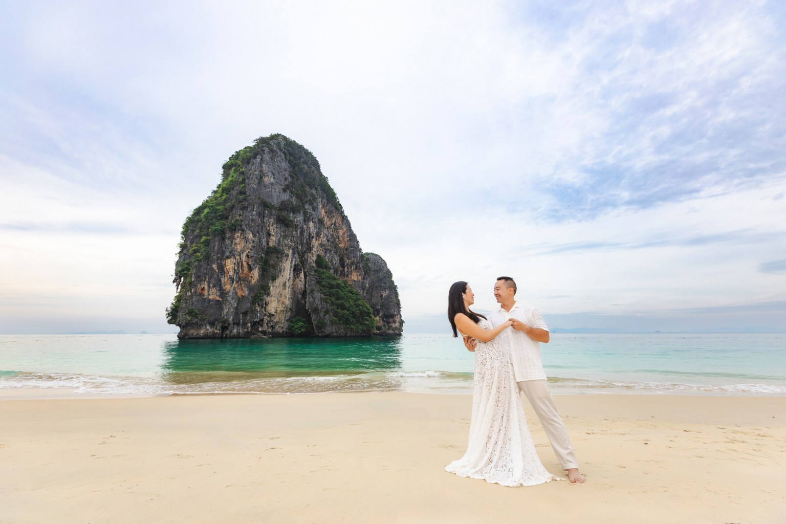 couple in wedding attire embracing on a beach with a large rock formation in the background