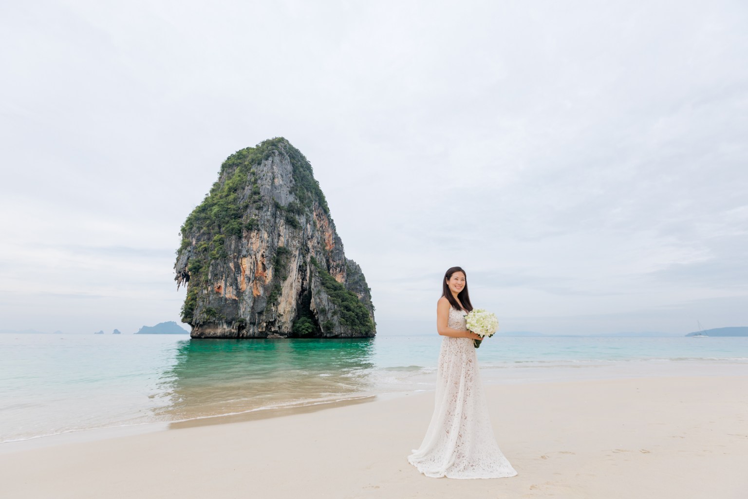 A woman in a white dress holds a bouquet while standing on a beach with a large rock formation in the background and calm blue waters.