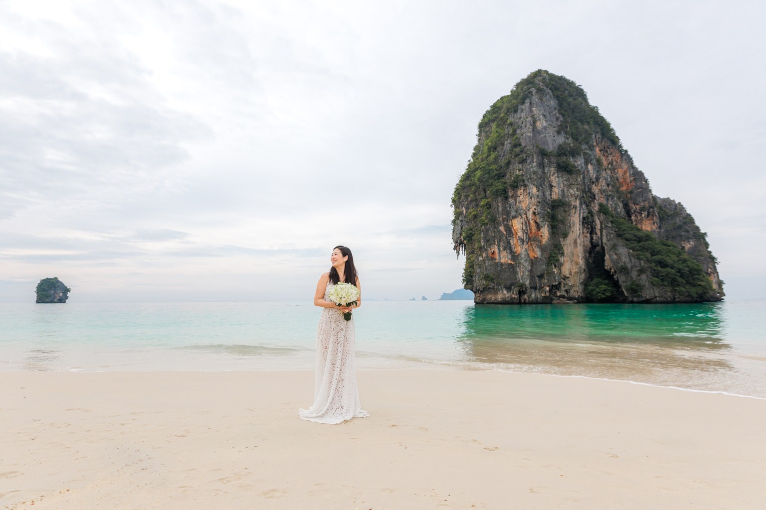 A woman in a lace dress stands on the beach holding a bouquet of flowers, with a large rock formation and calm turquoise water in the background.