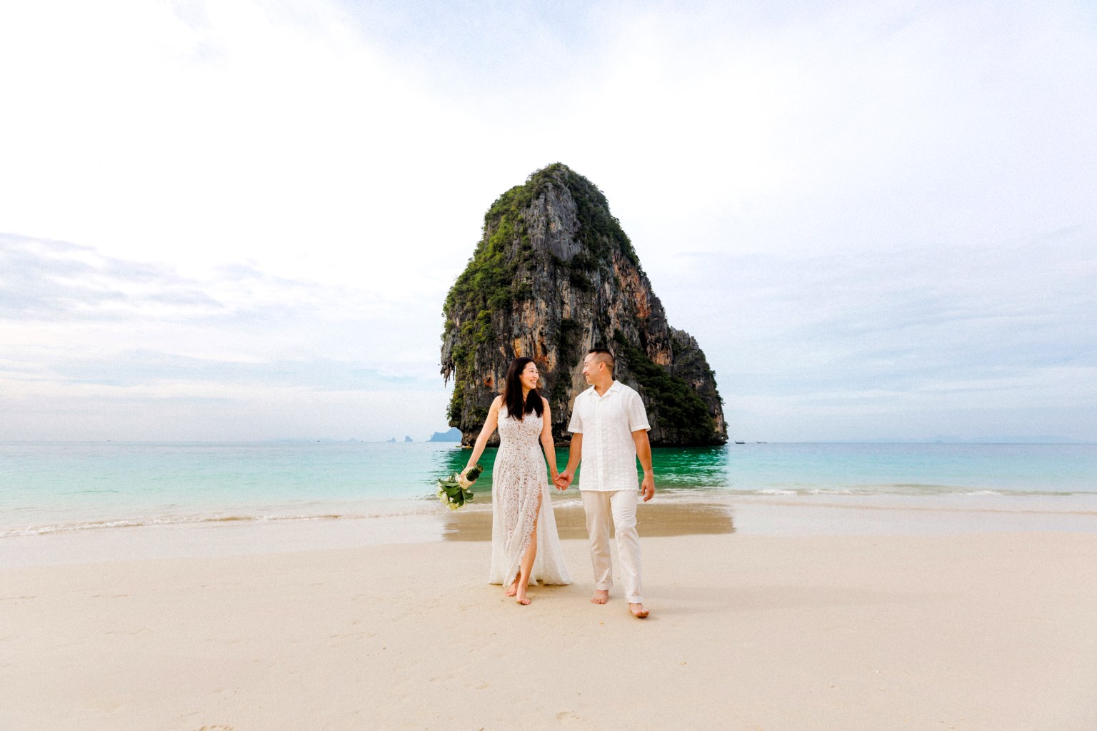 A couple walking hand-in-hand on a beach in Krabi, Thailand, with a large rock formation in the background and turquoise waters.