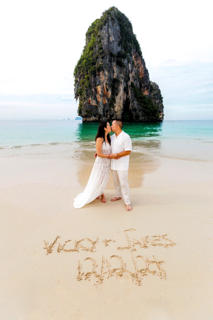 Couple kissing on a beach in front of a rock formation, with their names written in the sand.