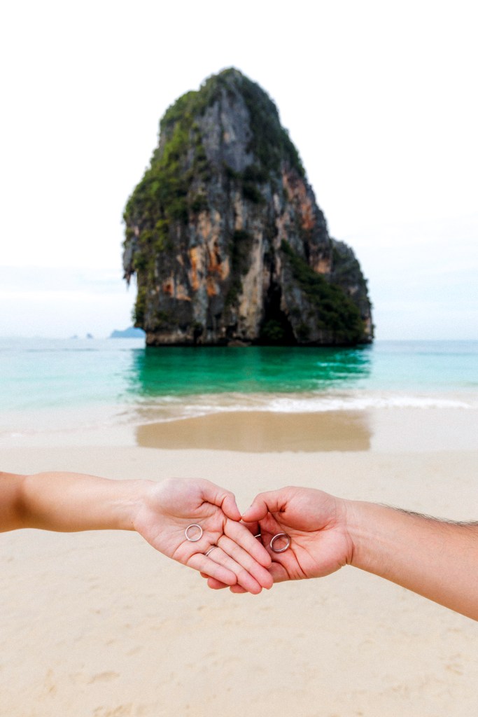 A close-up of a couple's hands holding wedding rings in front of a scenic beach with a large rock formation in the background.