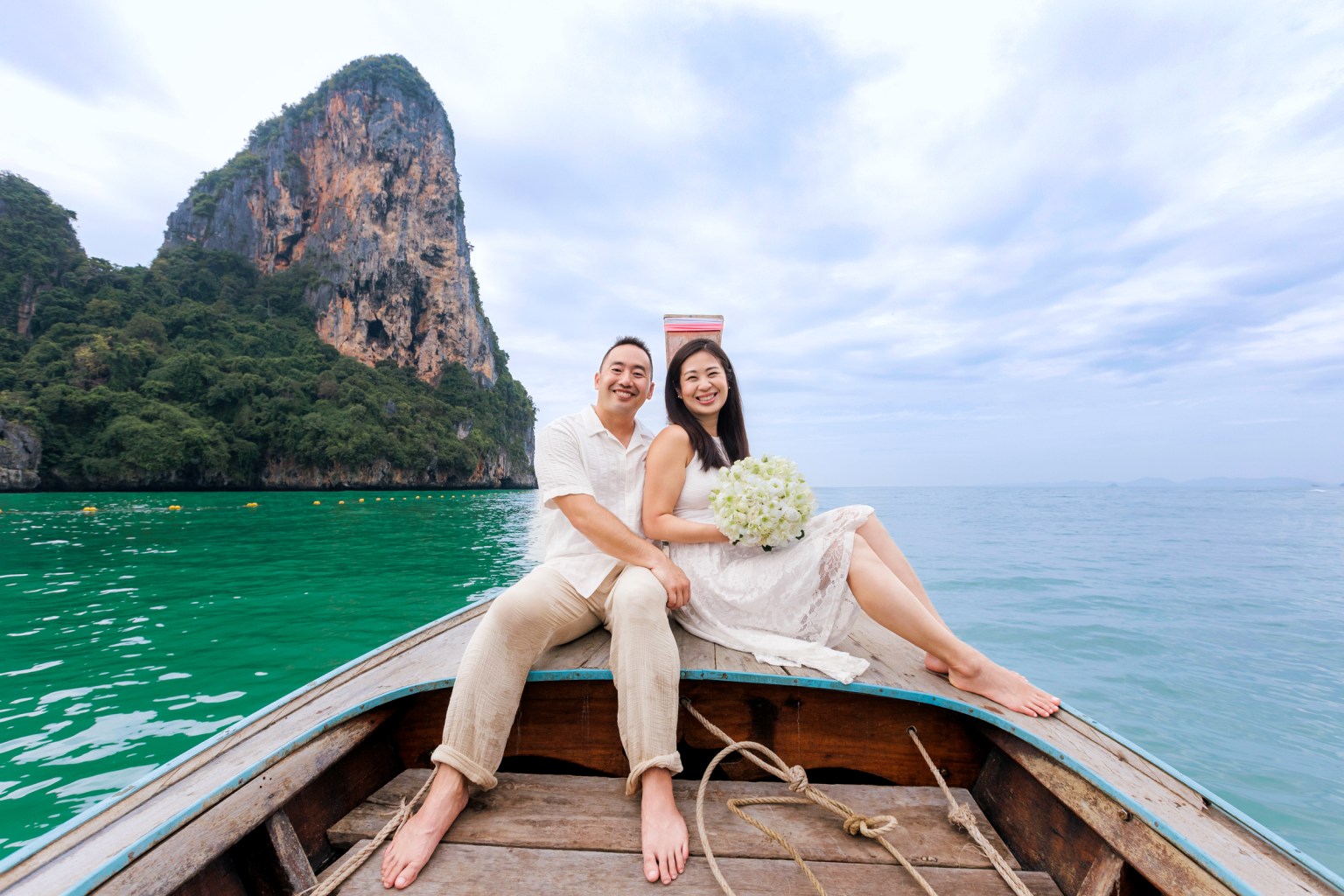 Honeymoon couple sitting on a long tail boat, smiling, with a bouquet of flowers and a scenic view of green waters and rocky cliffs in the background.