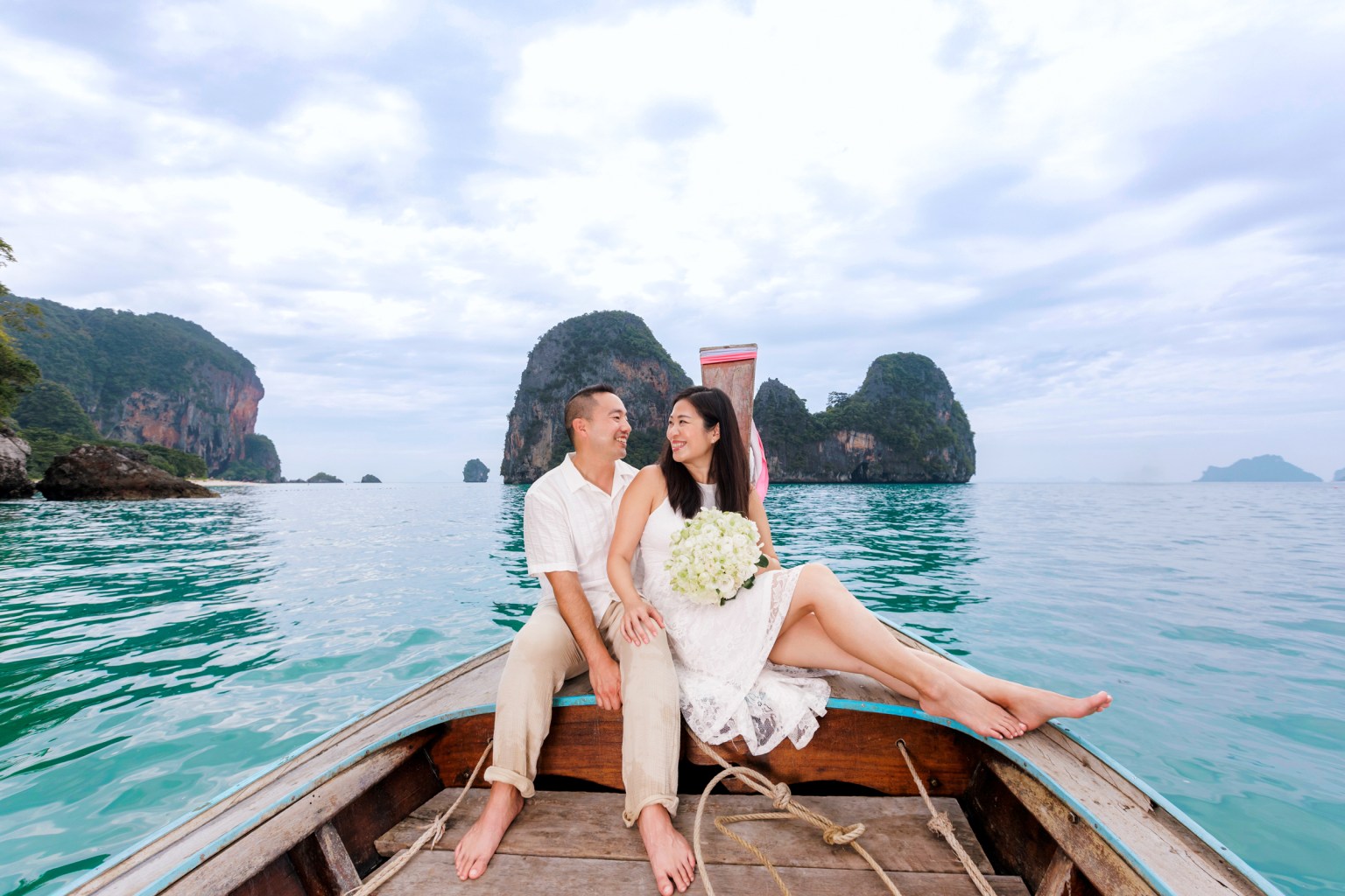 A couple sitting on a long tail boat, smiling at each other, surrounded by scenic cliffs and clear turquoise water.