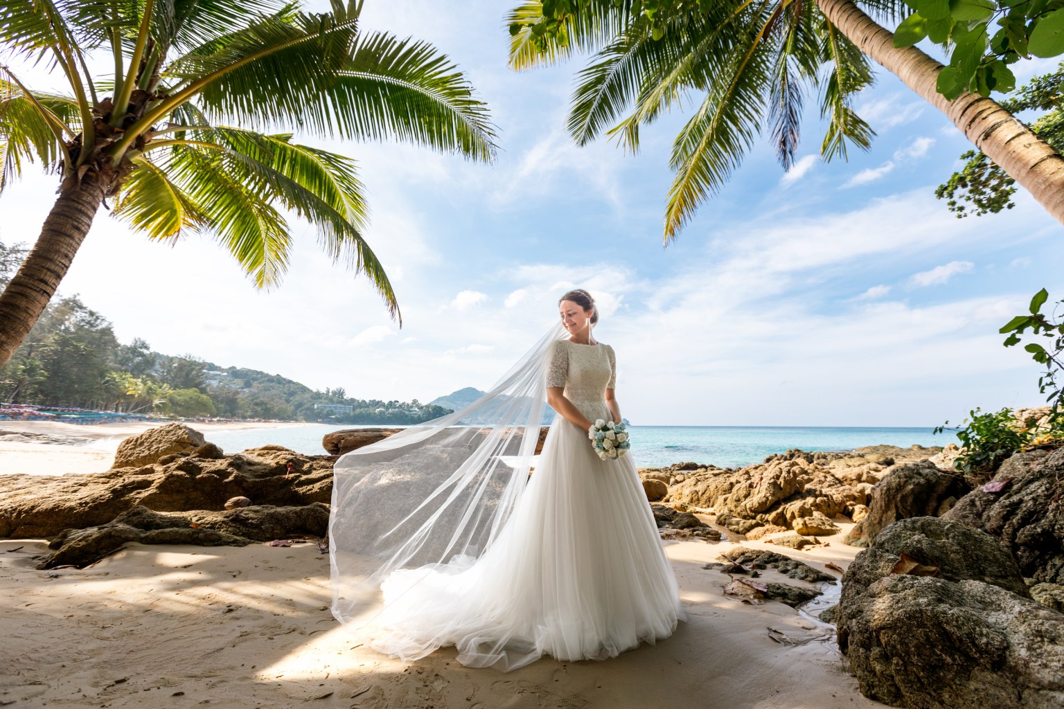 Bride in a wedding dress holding a bouquet, standing on a beach with palm trees and ocean in the background at surin beach phuket.