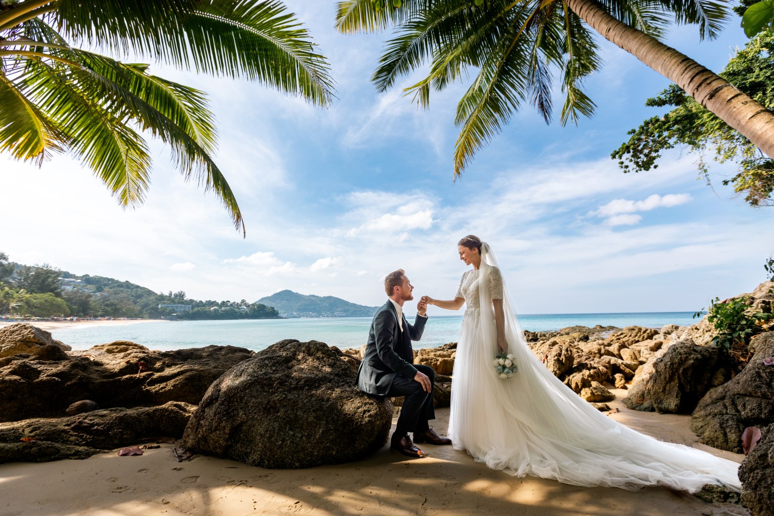 A couple engaged in a romantic moment at Surin beach, Phuket, with the bride in a flowing white wedding dress, holding a bouquet and smiling at her partner who is seated on a rock.