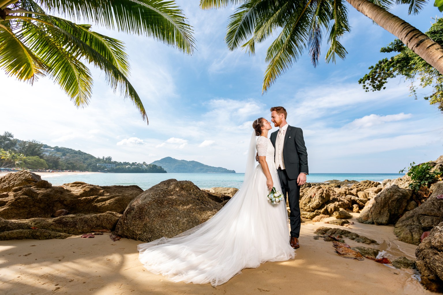 A bride and groom embracing on a beach, with palm trees in the foreground and a scenic ocean view behind them.