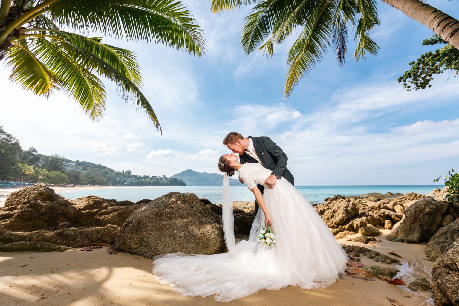 A couple sharing a romantic kiss on a beach surrounded by palm trees and rocky outcrops, with the ocean in the background.
