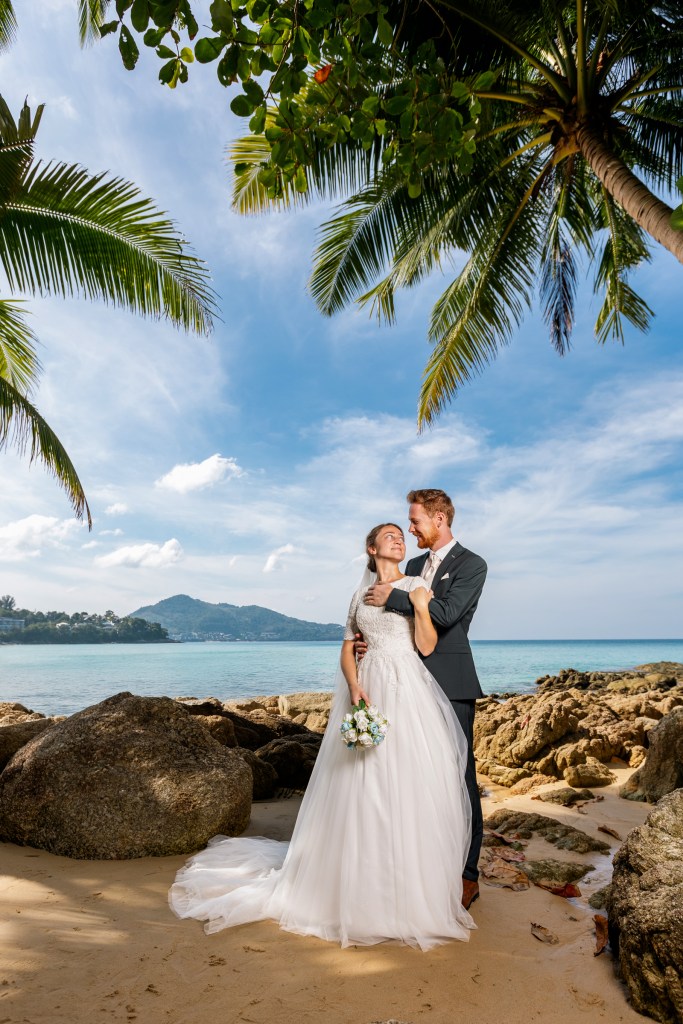 A bride and groom embrace on a sandy beach, surrounded by palm trees and a scenic ocean view in Phuket.