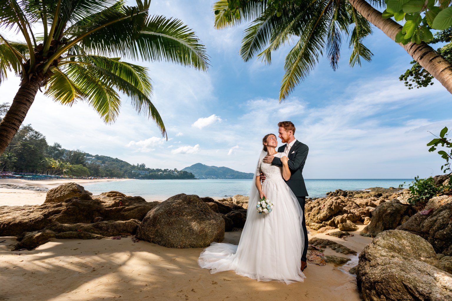 A couple in wedding attire smiling at each other on a beach with palm trees and rocky outcrops in the foreground, and the ocean in the background at surin beach phuket