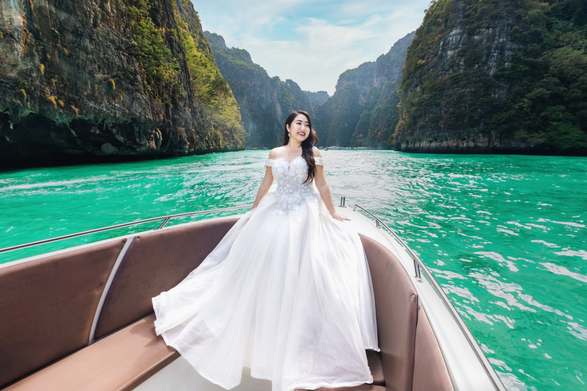 A bride in a white wedding dress poses on the bow of a boat surrounded by lush green cliffs and turquoise water.