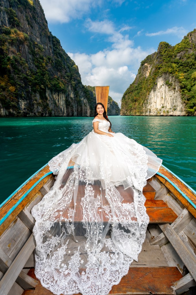 A bride in a white wedding dress sitting on a wooden boat at Pileh Lagoon, surrounded by towering cliffs and clear blue waters.