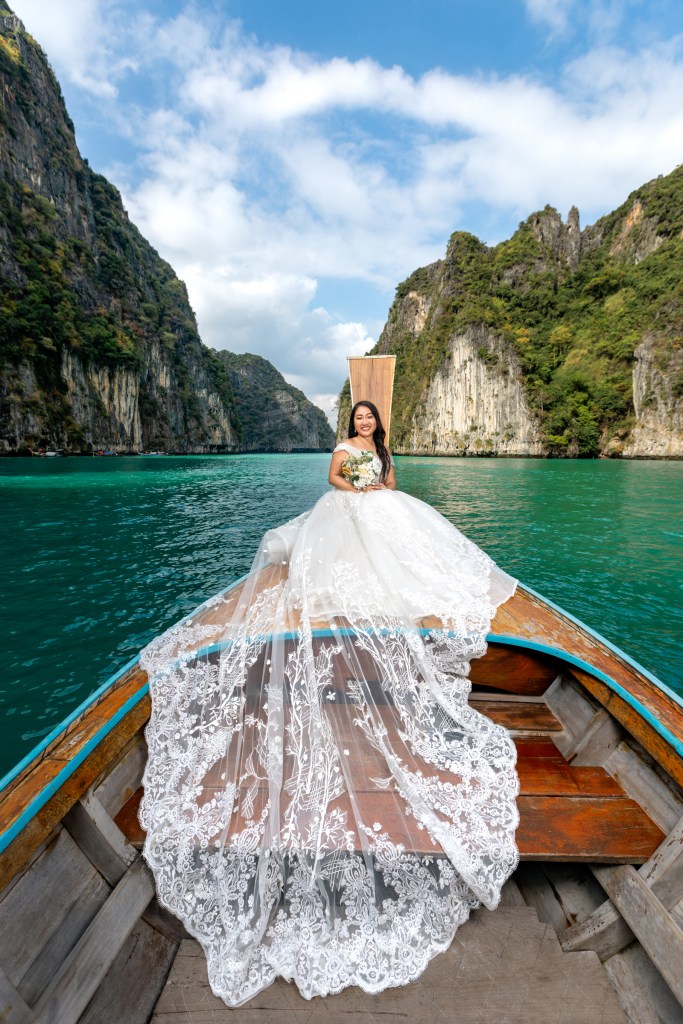 Bride in a wedding dress sitting on a long tail boat at Pileh Lagoon, Phi Phi Island, surrounded by steep cliffs and clear turquoise water.