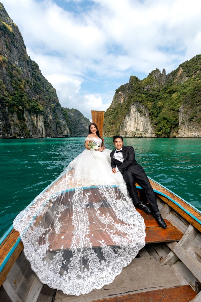 A couple in wedding attire poses on a long-tail boat at Pileh Lagoon, Phi Phi Island, with lush green cliffs in the background and a clear blue sky.