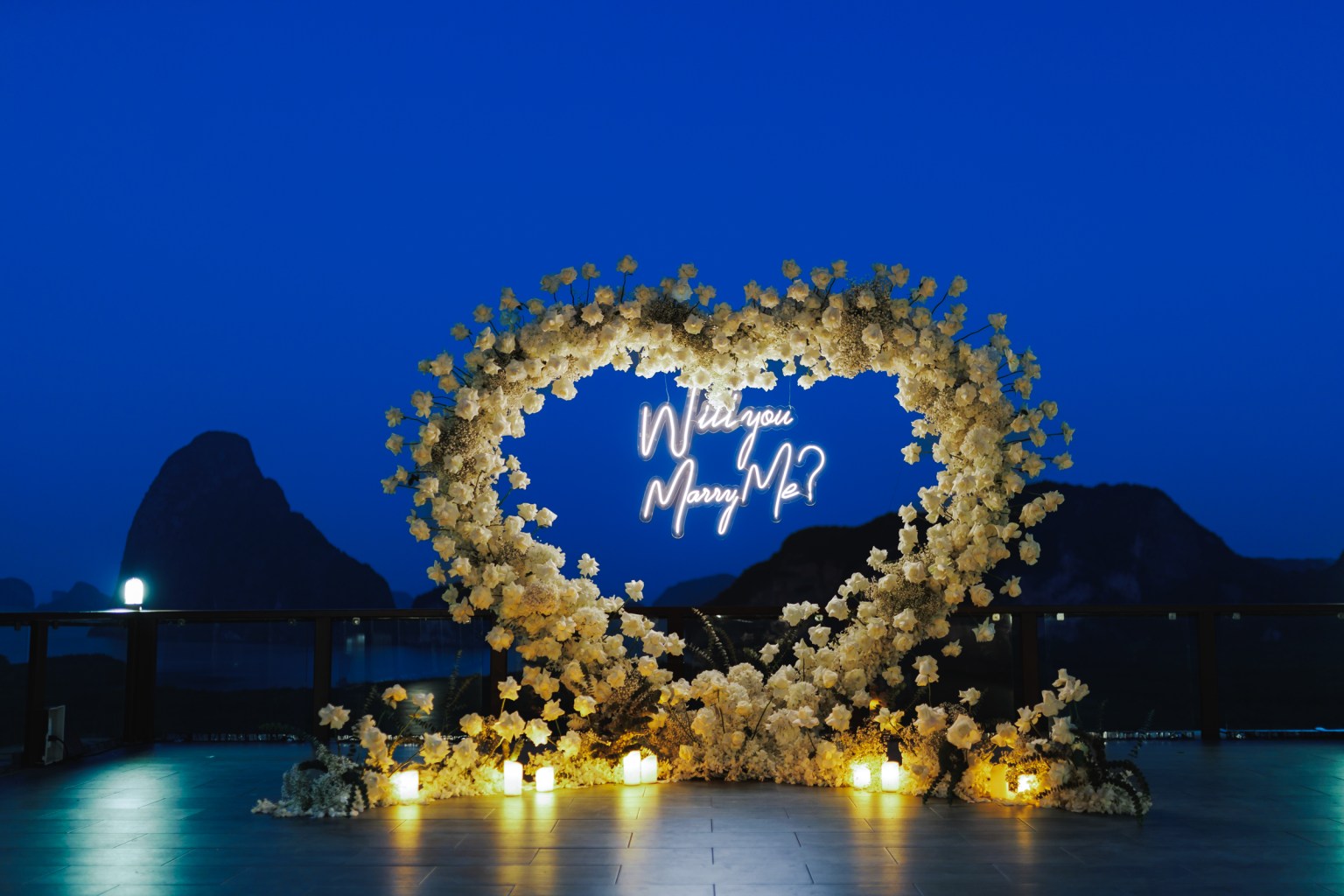 Wedding proposal setup with a neon sign saying 'Will you Marry Me?' surrounded by white flowers and candles, set against a twilight sky at samet nangshe 