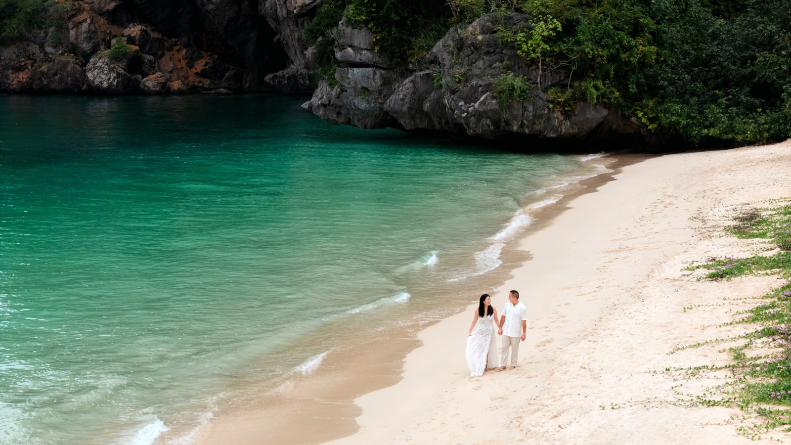 A couple walking hand in hand along a picturesque beach, with clear turquoise water and rocky cliffs in the background, capturing a romantic moment.