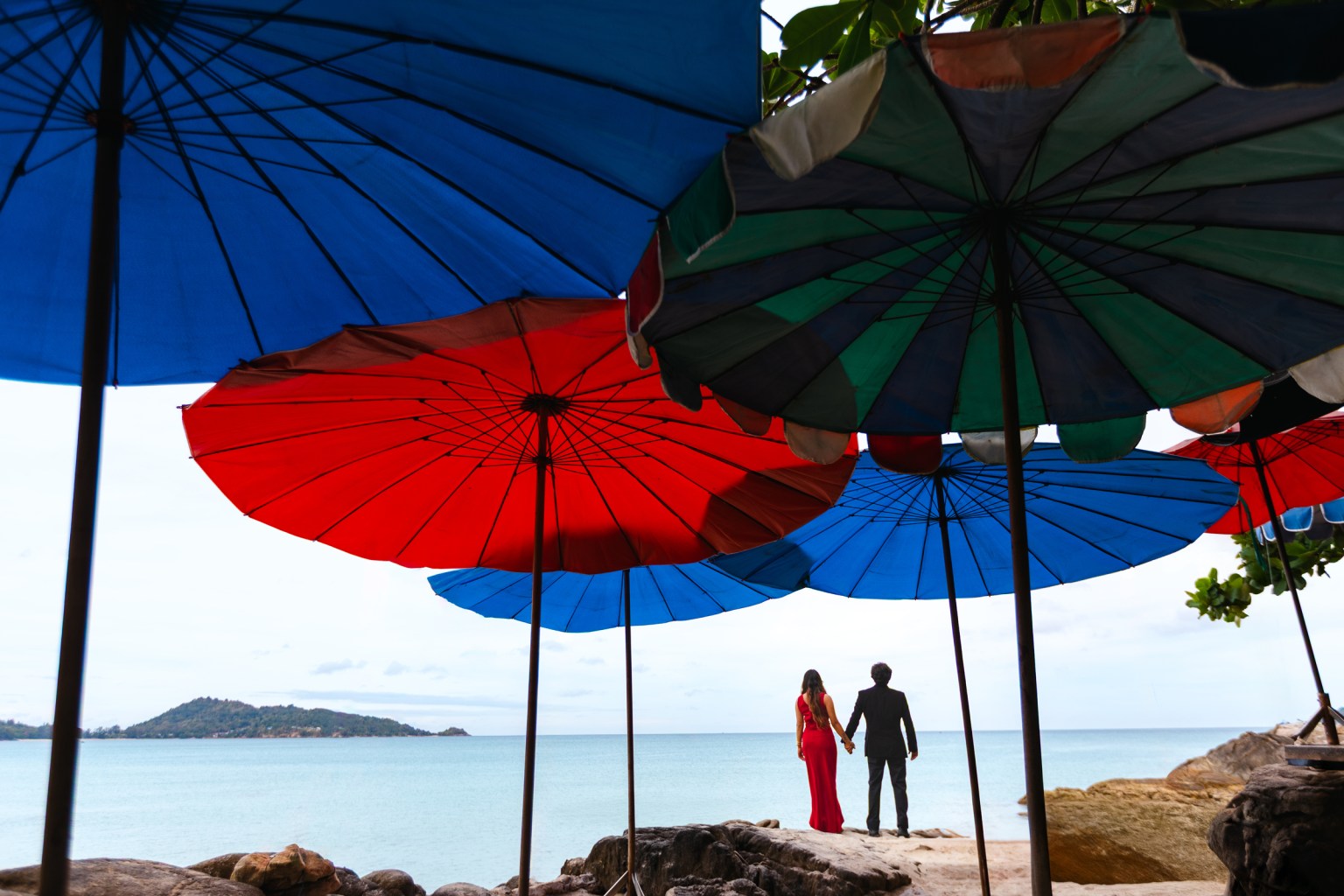 A couple holding hands under colorful beach umbrellas, looking out at the sea.