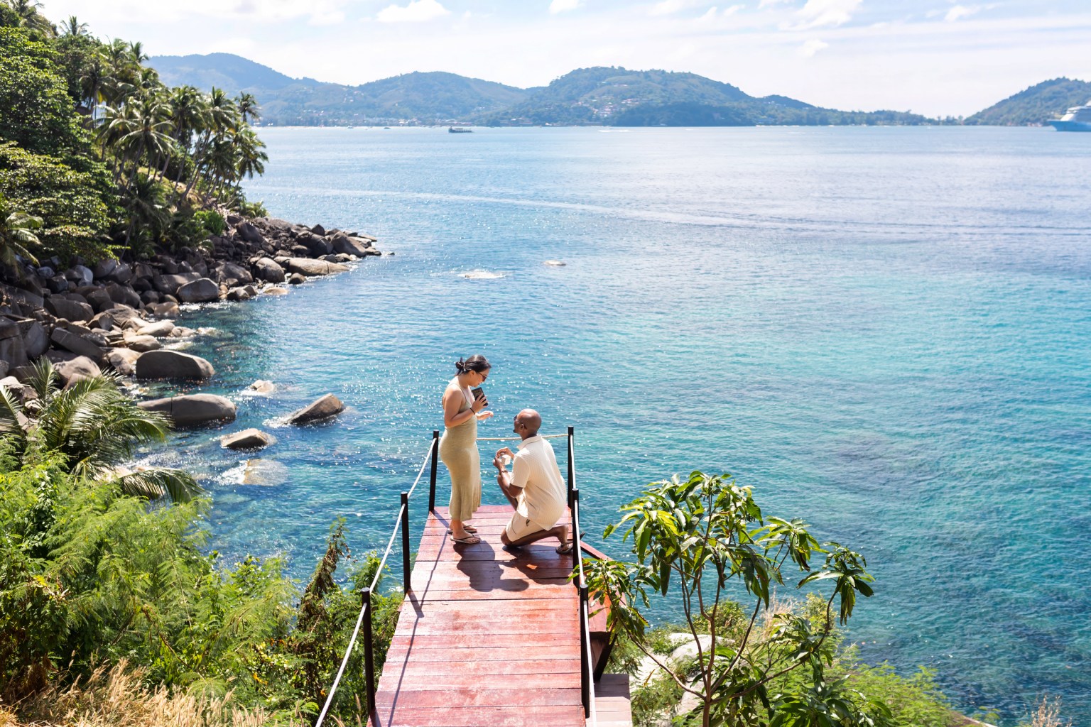 A couple at a marriage proposal on a wooden platform by the sea with turquoise water and mountains in the background.