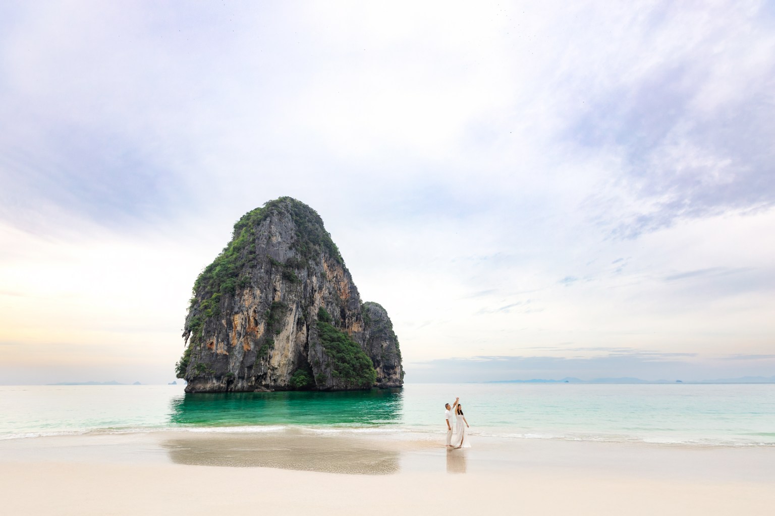 A couple in wedding attire dances on a serene beach at Phra nang cave beach in railay Krabi, Thailand, with a dramatic limestone cliff in the background and clear turquoise waters.