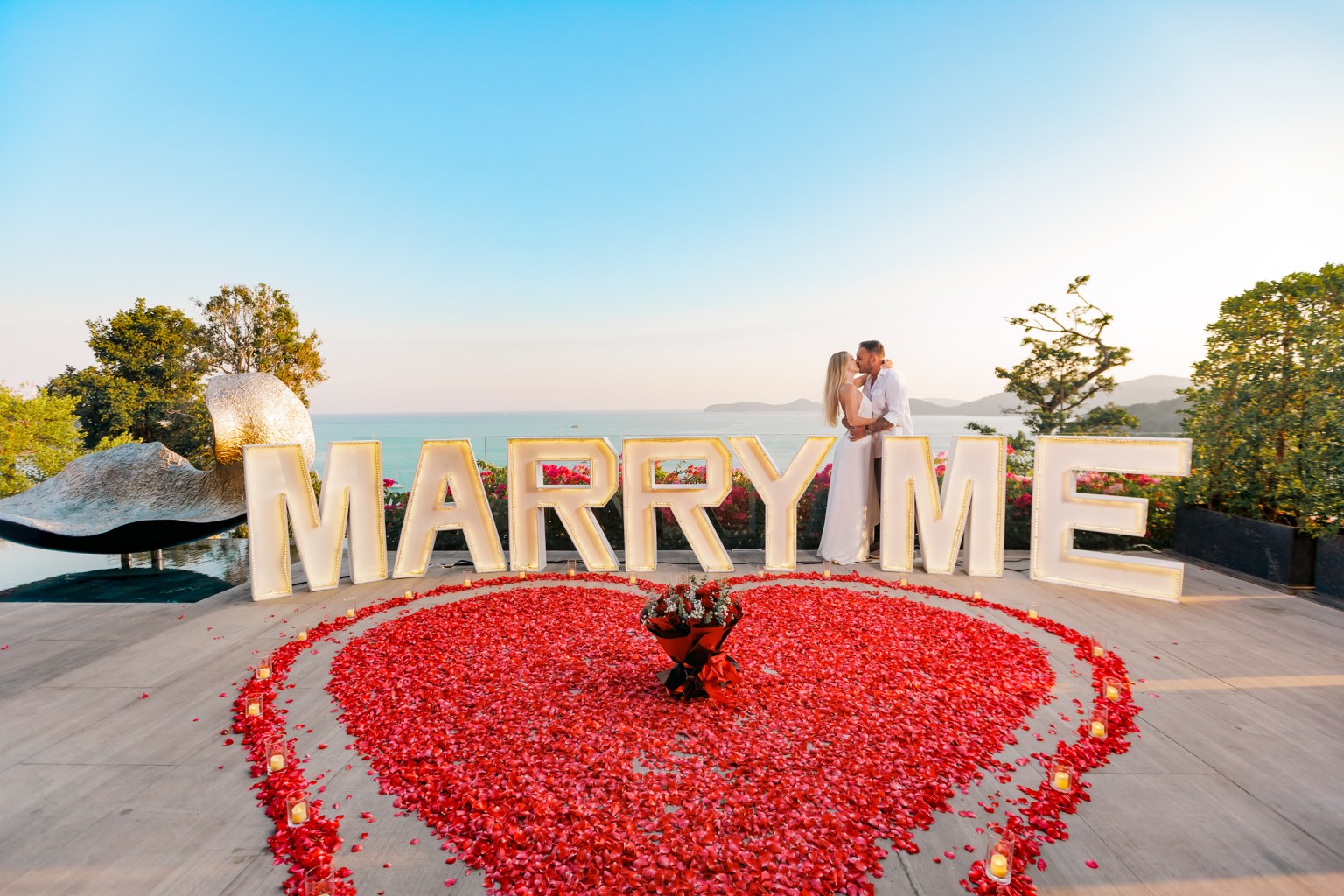 Couple kissing in front of a 'MARRY ME' display surrounded by rose petals and decorative lights.