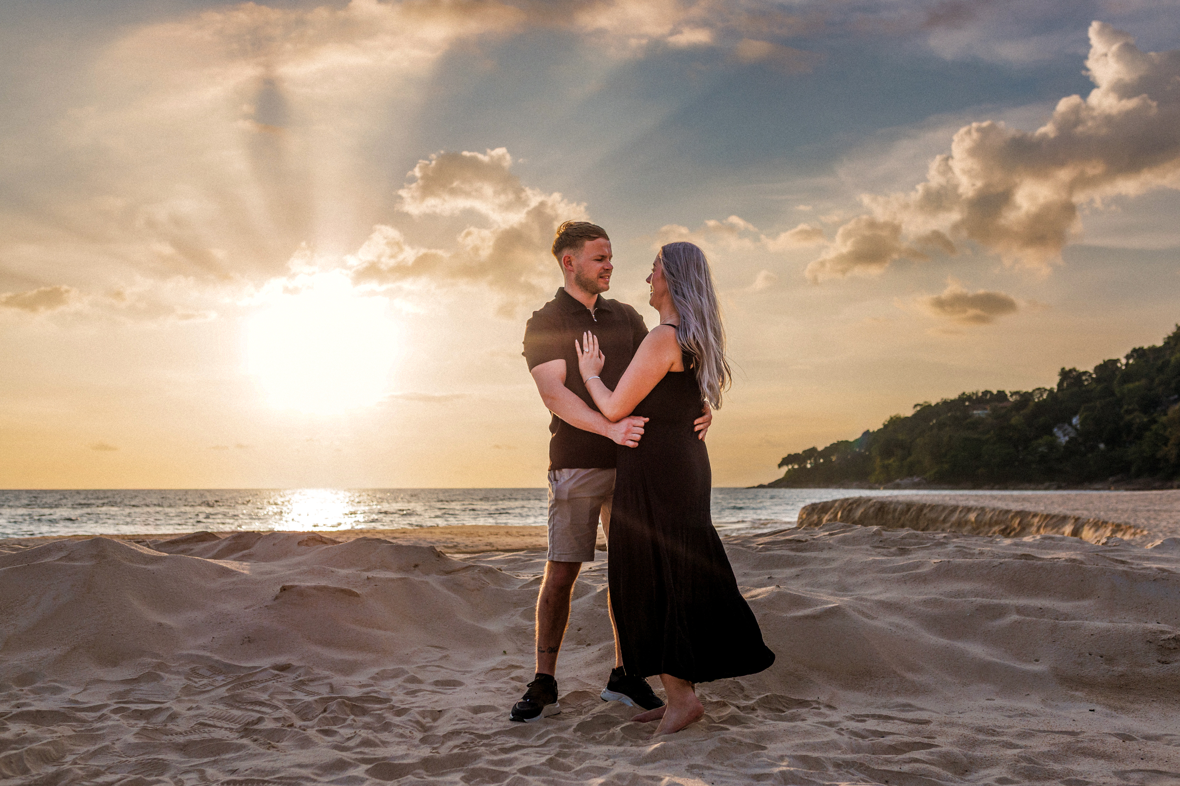 A couple stands on a sandy beach at sunset, embracing each other with the ocean in the background and rays of sunlight shining through the clouds.