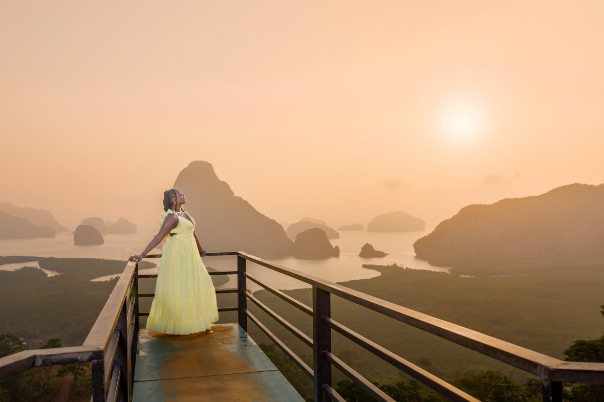 A woman in a flowing yellow dress standing on a viewpoint balcony overlooking a misty landscape of hills and water at sunrise.